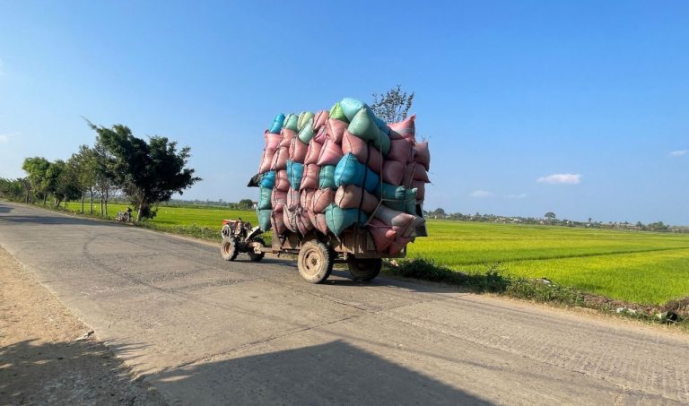 A heavily loaded cart stacked with colorful fabric bundles travels along a rural road past green rice paddies.