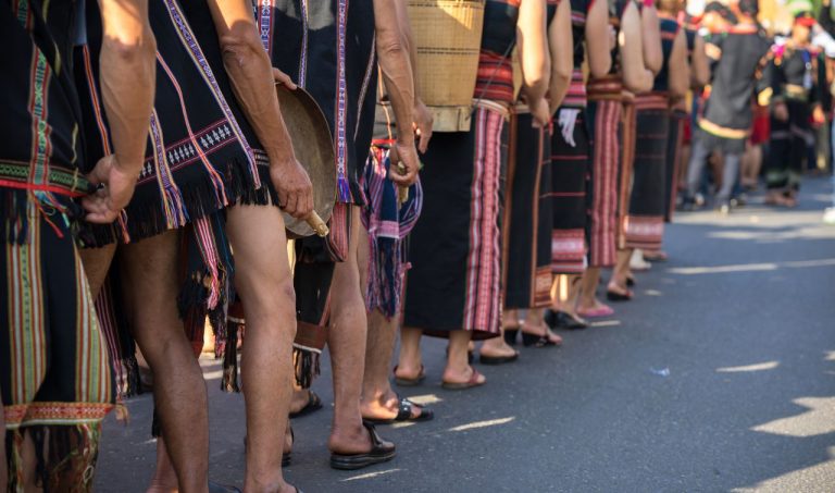 People wearing traditional woven textiles with colorful striped patterns stand in a line during a daytime celebration.