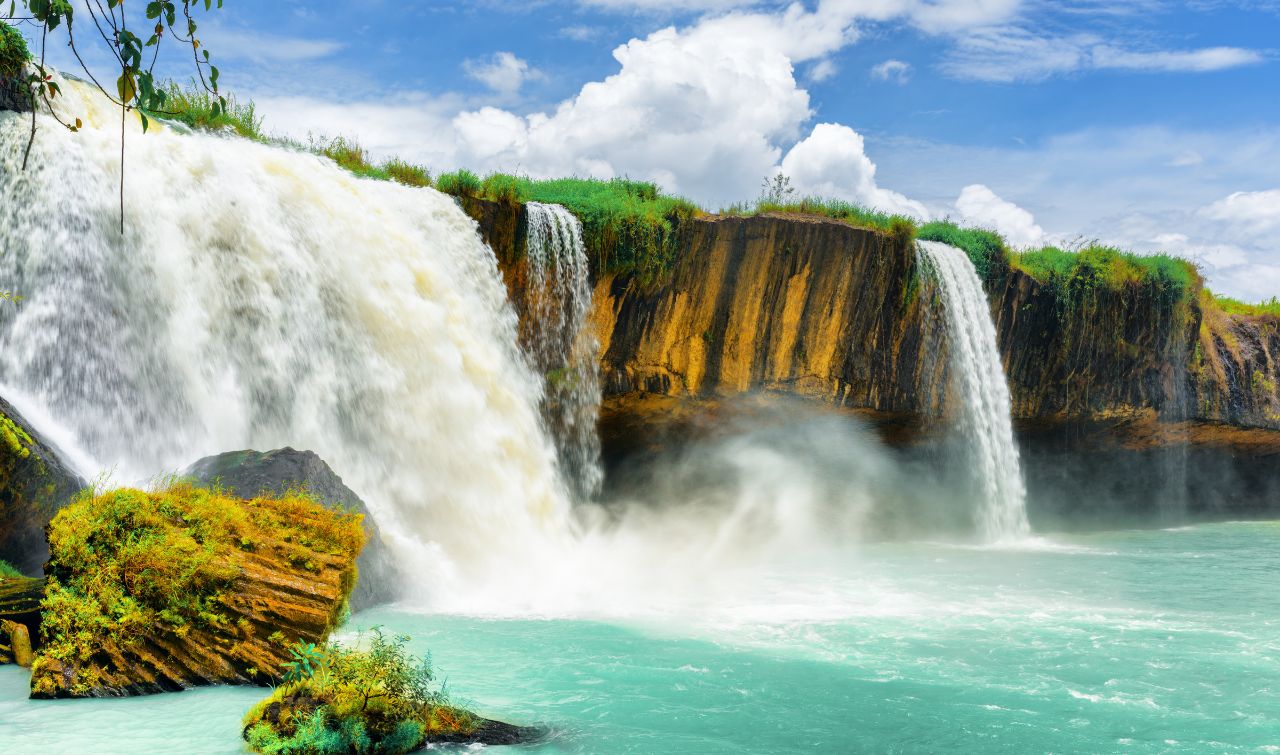 Powerful waterfall cascades into turquoise pool with moss-covered rocks and lush green cliff top.